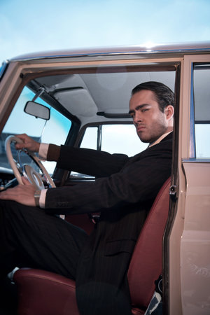 Retro 60s fashion business man wearing grey suit with tie sitting in classic car.