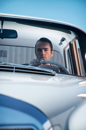 Retro 60s fashion business man wearing grey suit with tie sitting in classic car.
