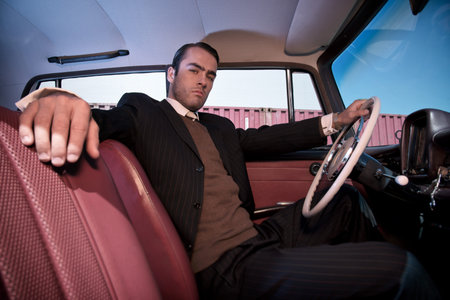 Retro 60s fashion business man wearing grey suit with tie sitting in classic car.
