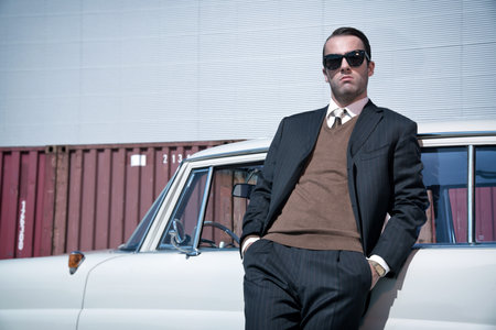 Retro 60s fashion business man wearing grey suit and black sunglasses standing against classic car.