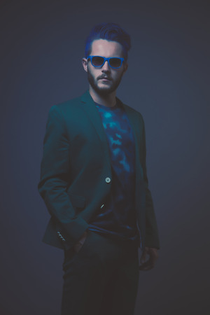 Man with blue hair and brown beard wearing green suit. Wearing blue sunglasses. Male fashion. Studio shot against dark background.の写真素材