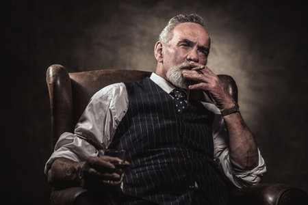 In chair sitting senior business man with cigar and whisky. Gray hair and beard wearing blue striped...