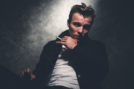 Cigarette smoking retro fifties cool rebellion fashion man wearing white shirt and jeans jacket. Gray wall.の写真素材
