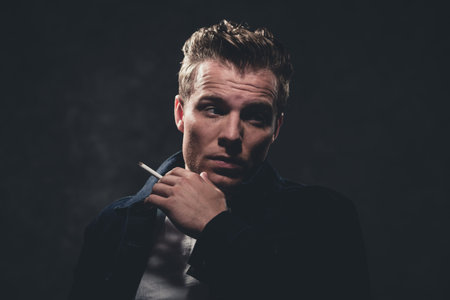 Cigarette smoking retro fifties cool rebellion fashion man wearing white shirt and jeans jacket. Gray wall.の写真素材