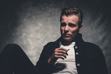 Cigarette smoking retro fifties cool rebellion fashion man wearing white shirt and jeans jacket. Gray wall.の写真素材