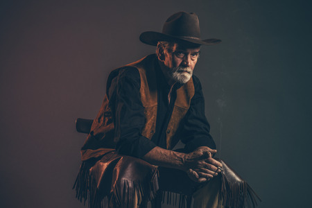 Cigarette smoking old rough western cowboy with gray beard and brown hat. Low key studio shot.の写真素材