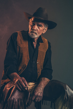 Cigarette smoking old rough western cowboy with gray beard and brown hat. Low key studio shot.の写真素材
