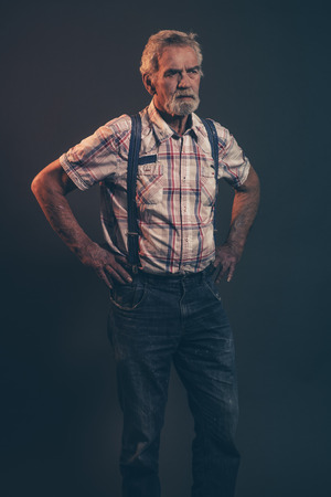 Characteristic senior man with gray hair and beard wearing checkered shirt with braces and blue jeans. Low key studio shot.の写真素材
