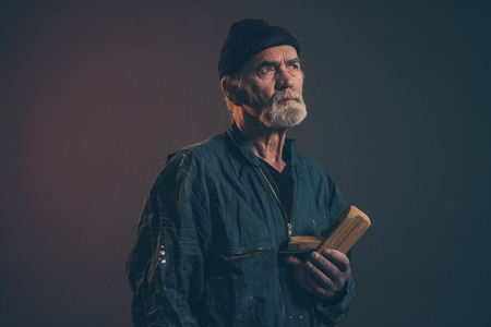 Senior carpenter with gray hair and beard holding old plane wearing black hat with green overalls. Low key studio shot.の写真素材
