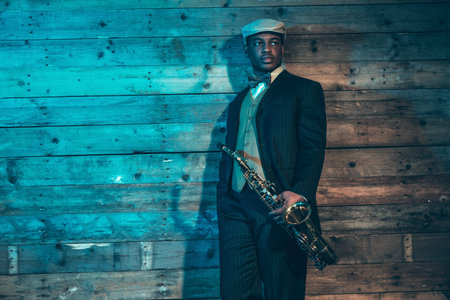 Vintage african american jazz musician with saxophone in front of old wooden wall. Wearing suit and cap.の写真素材