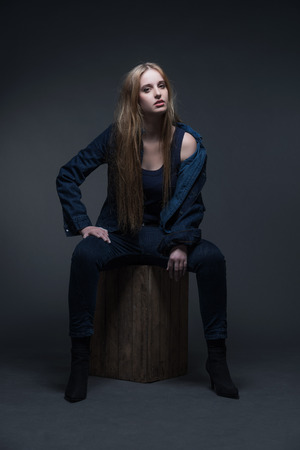 Woman jeans winter fashion. Sitting on vintage wooden box. Long blonde hair. Studio shot against grey.の写真素材