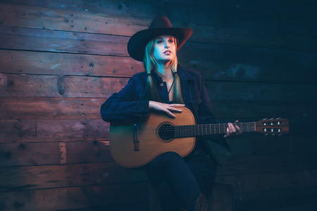 Cowgirl country singer with acoustic guitar. Wearing blue jeans and brown hat. In front of wooden wall.の写真素材