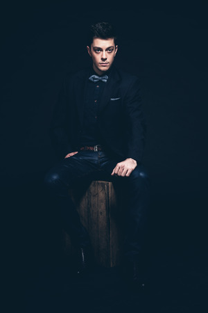 Retro fashion man with blue shirt, jacket, jeans and bow tie. Sitting on vintage wooden box. Short dark hair. Studio shot against black.の写真素材