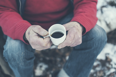 Hands of a senior man sitting outdoors in winter snow drinking a mug of freshly brewed black coffeeの写真素材