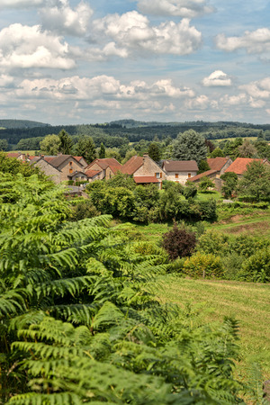 Quaint rural stone village in lush countryside viewed across the top of leafy shrubs under a blue sky with pretty white cloudsの写真素材