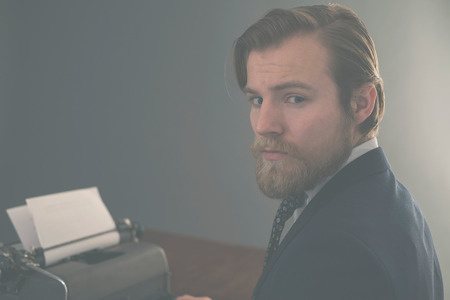 Handsome bearded vintage businessman sitting at his desk with an old manual typewriter turning to look thoughtfully at the camera, aged effectの写真素材