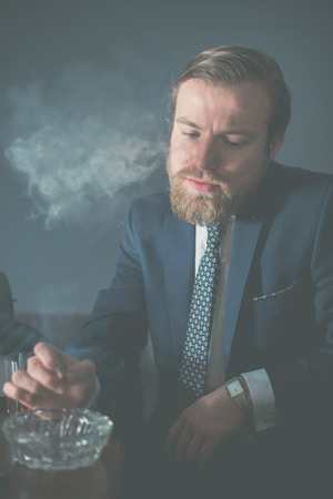 Handsome bearded businessman sitting smoking knocking off ash from his cigarette into an ashtray in a smoke laden roomの写真素材