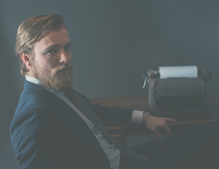 Handsome old-fashioned businessman sitting at his desk in front of a vintage manual typewriter turning to look at the camera, aged effectの写真素材