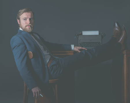 Successful vintage businessman relaxing in the office with his feet on the desk alongside an old manual typewriter looking thoughtfully to the side with a pensive expressionの写真素材