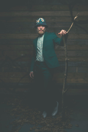 Stylish Middle Age Man in Fashionable Green Outfit with Hat Leaning Againts on a Dry Tree Branch in Front of a Rustic Wall Background.の写真素材