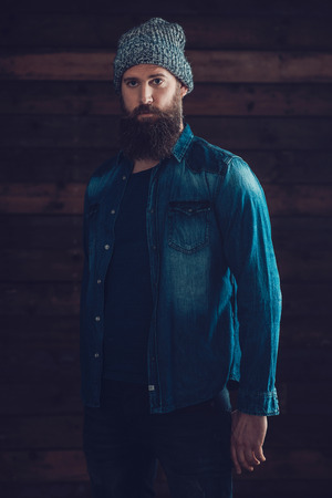 Serious Young Man with Long Beard Wearing Casual Denim Jacket and Knitted Hat Outfit, Standing In Front a Wooden Wall Background.の写真素材