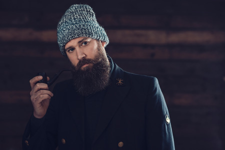 Close up Gorgeous Man with Long Beard, Wearing Black Coat and Gray Bonnet, Holding a Smoking Pipe While looking at the Camera.の写真素材