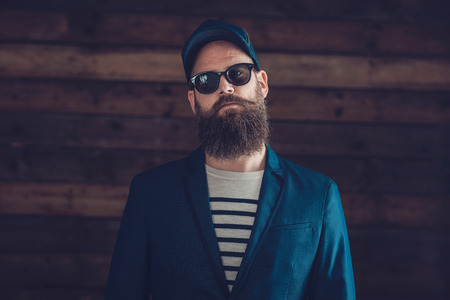 Close up Stylish Goatee Man, in Dark Blue Formal Coat with Cap and Sunglasses Facing at the Camera on a Wooden Wall Background.の写真素材