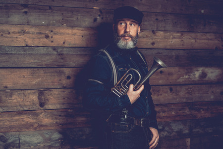 Half Body Shot of a Serious Middle Age Man in Black Attire with Cap Holding a Trumpet Instrument in front Wooden Wall While Looking to the Left of the Frame.の写真素材