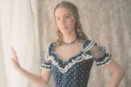 Faded aged portrait of a pretty blond girl in vintage fashion standing with her hand to a filmy white curtain looking at the camera with a serious expressionの写真素材