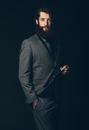Portrait of an Attractive Young Guy with Beard and Mustache, Wearing Classy Formal Suit, Holding his Eye Wear with the Other Hand in his Pocket. Isolated on Black.の写真素材