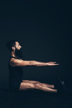 Athletic Young Man with Goatee Beard Sitting on the Floor with Both Arms and Legs Stretched Forward, Isolated on Black Background.の写真素材