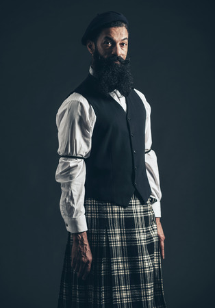 Attractive bearded man wearing a plaid Scottish kilt and waistcoat standing looking at the camera wuith a serious expression over a dark backgroundの写真素材