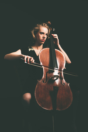 Attractive young woman playing the cello in the darkness at a recital or classical concert, full length portraitの写真素材