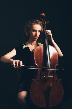 Young woman playing the cello in a classical concert or recital sitting in the darkness with a serious expression as she awaits her cue to start playingの写真素材