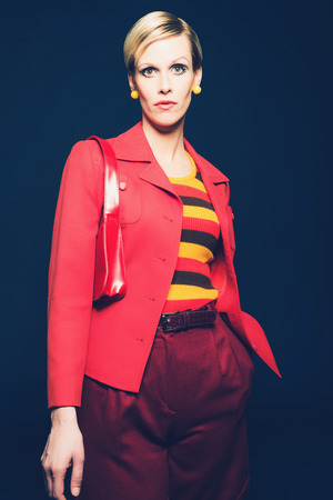 Portrait of a Stylish Blond Woman in Red and Yellow Fashion, Wearing Striped Shirt, Red Blazer and Shorts with Shoulder Bag, Looking at the Camera. Isolated on Dark Blue.の写真素材