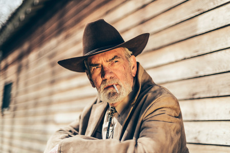 Close up Old Western Man Smoking a Cigarette While Sitting on Steps In Front of his house and Looking Afar Seriously.の写真素材