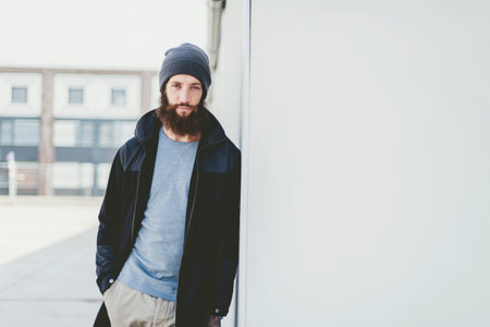 Serious Young Man in Winter Outfit Leaning on White Building Wall and Looking at the Camera.