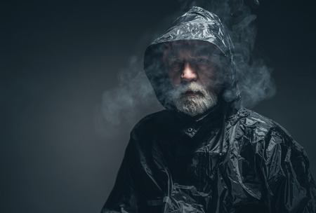 Close up Pensive Old Bearded Guy in Black Rain Jacket with Cigarette Smoke Against Black Background.の写真素材