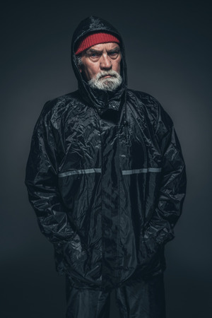 Portrait of a Serious Adult Guy with White Facial Hair, Wearing Black Winter Clothing, Looking Straight at the Camera Against Black Background.の写真素材