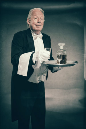 Senior Male Butler Wearing Tuxedo and Serving from Tray with Liquor Bottle and Crystal Cordial Glassesの写真素材