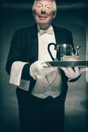 Portrait of Friendly Looking Senior Male Butler Wearing Tuxedo and Holding Tray with Tea Service for One with Pot and Cupの写真素材
