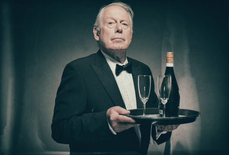 Waist Up Portrait of Professional Looking Senior Male Butler Wearing Suit with Bow Tie and Carrying Tray with Bottle of Champagne and Two Glass Flutesの写真素材