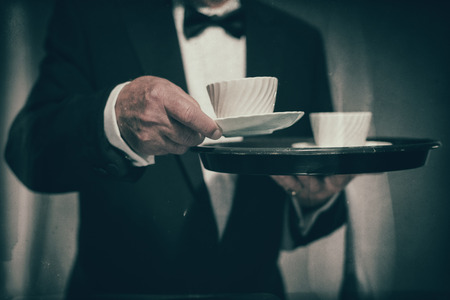 Close Up of Male Butler Wearing Formal Suit and Bow Tie Carrying Tray of White Coffee Mugs and Serving Cup Towards Cameraの写真素材