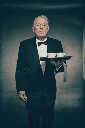 Senior Male Butler Wearing Formal Suit and Bow Tie Holding Tray with Two White Coffee Cups in Dimly Lit Studioの写真素材