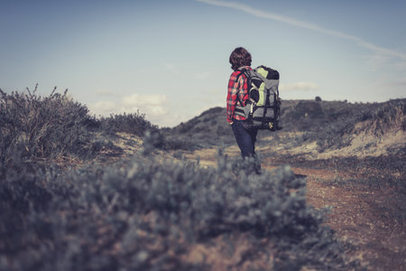 Backpacker wearing a large backpack of camping gear walking through hilly scrub standing facing away...