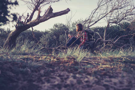 Outdoors Man with Beard and Long Hair Wearing Backpack Sitting on Fallen Tree in Forest Clearing, Man Taking a Rest from Hiking and Looking into the Distanceの写真素材