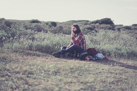 Backpacker taking a rest in the wilderness sitting with his backpack on the ground relaxing and staring thoughtfully into the distanceの写真素材