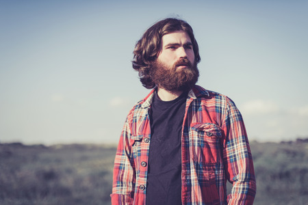 Handsome bearded man wearing a red plaid shirt standing outdoors in nature looking off to the right of the frame with a thoughtful expressionの写真素材
