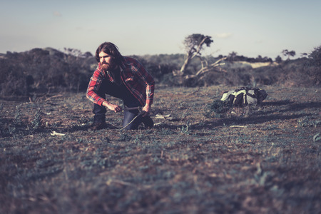 Hiker collecting firewood to make a campfire to cook a meal leaning down to pick up dry branches with his backpack on the ground behind himの写真素材