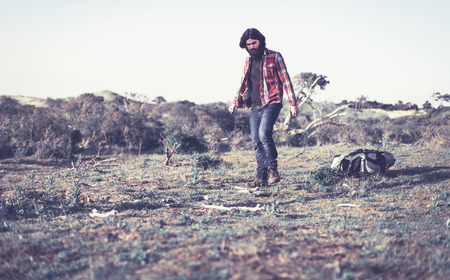 Hiker collecting firewood to make a campfire to cook a meal leaning down to pick up dry branches with his backpack on the ground behind himの写真素材